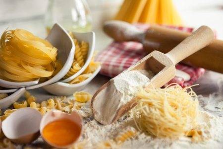 Various pasta, wooden scoop with flour and eggs on floured surfaceの写真素材