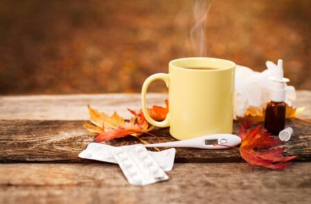 Cup with hot tea and thermometer showing high temperature along with pills and nose drops on wooden surface, flu season in autumnの写真素材