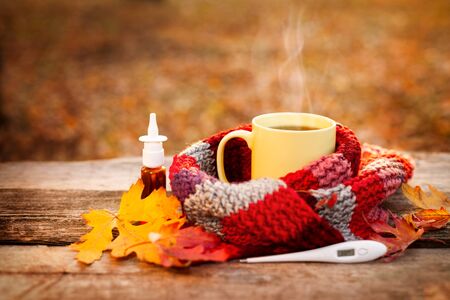 Tea mug and warm scarf with nose drops and fallen leaves on wooden surfaceの写真素材