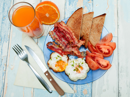 Fried eggs with toast, bacon and tomato on a plate, orange juice on the sideの写真素材