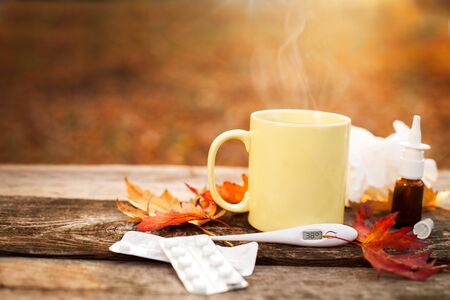 Mug with hot tea and thermometer showing high temperature along with pills and nose drops on wooden surface, flu season in autumnの写真素材