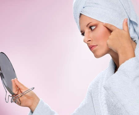 Young woman in bathrobe holding mirror and looking at her skinの写真素材