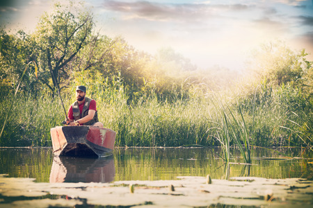 Fisherman with fishing rods is fishing in a wooden boat against background of beautiful nature and lake or river. Camping tourism relax trip active lifestyle adventure conceptの写真素材