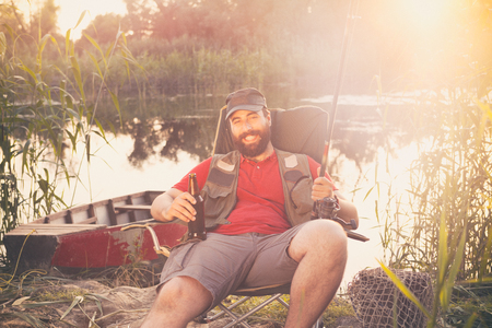 Smiling fisherman sitting with fishing rod and beer, leisure and hobbyの写真素材