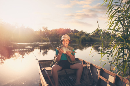 Smiling young fisher boy showing his first fish catch, happy boy sitting in boatの写真素材