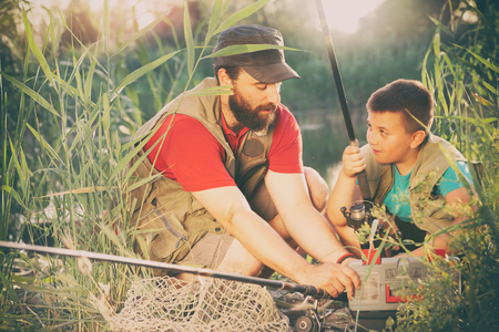 young father teaching his son to make a fishing rod, they are in grass on the shores of the lakeの写真素材
