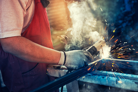 Strong man is a welder in T-shirt in a construction gloves, a metal product is welded with a welding machine in the garage, orange sparks fly to the sidesの写真素材