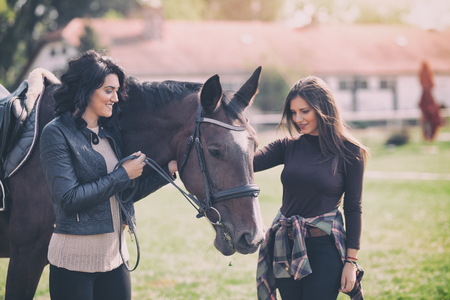 Two pretty woman enjoying horse company,  trendy  ladies at countryside outdoor, inspires hobby and love of horsesの写真素材