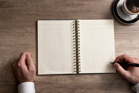 Direct view at hands writing on a blank paper. Office wooden desk with coffee, notebook, pencils. Copy space.の写真素材