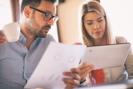 Young man and woman discussing work over a cup of coffee sitting in office. Businessman holding a paper in hand while talking to his business colleagueの写真素材