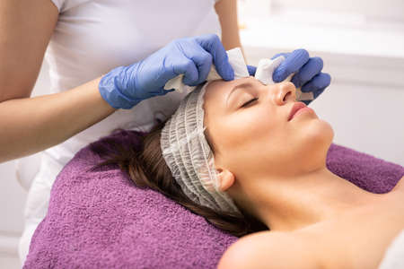 Beautician hands in rubber gloves wipe with cotton pad forehead of woman covered by cosmetic hat that lies on couch in beauty salonの写真素材