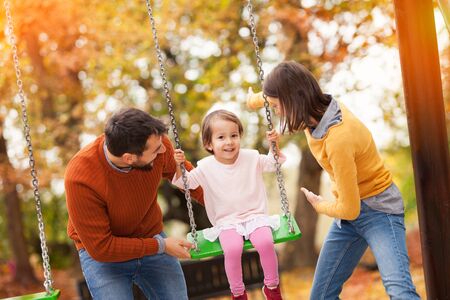 Family playing at a park together, having fun. The children, a 3 year old girl are on a swing set and their parents are pushing her swings.の写真素材