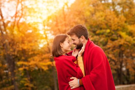 Loving couple. Beautiful young couple warped in blanket smiling and hugging  while standing  in autumn parkの写真素材