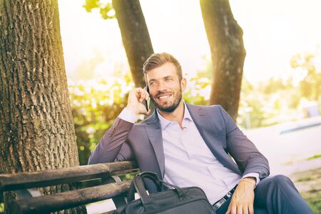 Businessman on park bench using mobile phone, telephone conversationの写真素材