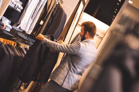 Young man choosing suit jacket during apparel shopping at clothing store