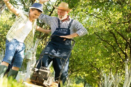 Grandfather and grandson together cutting grass with lawnmower in the orchard. Old and Young....