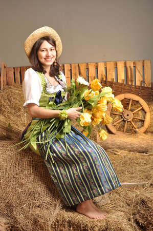Beautiful girl in dress sitting on hay with flowers in hand smilingの写真素材