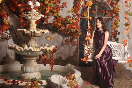 Beautiful girl in a purple dress sitting on a swing near the fountainの写真素材