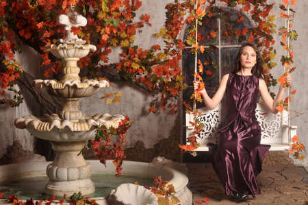Beautiful girl in a purple dress sitting on a swing near the fountainの写真素材