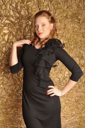 Portrait of a girl with red hair in a black dress on a gold background with hands on shoulders and hipsの写真素材