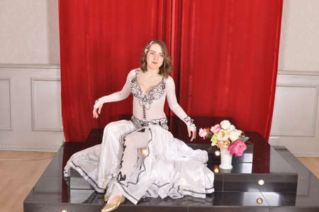 Beautiful girl in white oriental dance costume with flowers sitting on the podiumの写真素材
