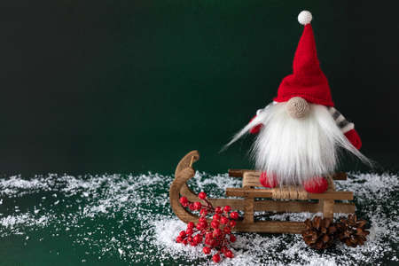 Scandinavian gnome with holly berry and pine sitting on wooden sledge on dark green background. New year greeting card designの写真素材