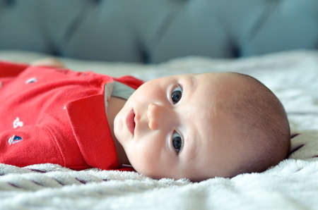 the baby is lying on the bed in a red T-shirt on a white striped blanket and looks at the camera. in the background, the headboard of the bed is gray in the out of focus.の写真素材