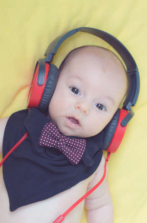 the baby in red headphones and a black bib with a burgundy bow tie with white polka dots looks at the camera .background yellow matte textileの写真素材