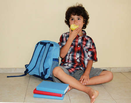 A beautiful smiling child with apple sitting on the floor surrounded with books and schoolbag