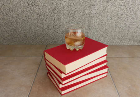 Glass of whiskey with ice on pile of red booksの写真素材
