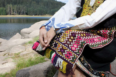 A young woman dressed in Bulgarian folklore costume is sitting near a lake, no faceの写真素材