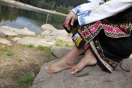 A young woman dressed in Bulgarian folklore costume is sitting near a lake, no faceの写真素材