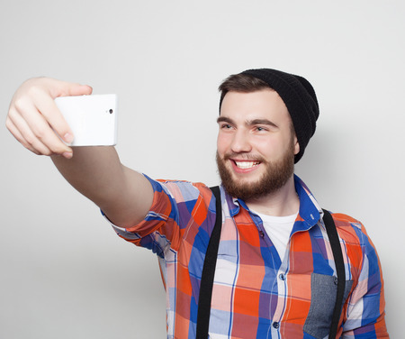 Life style concept: a young man with a beard  in shirt holding mobile phone and making photo of himself while standing against grey background.の写真素材
