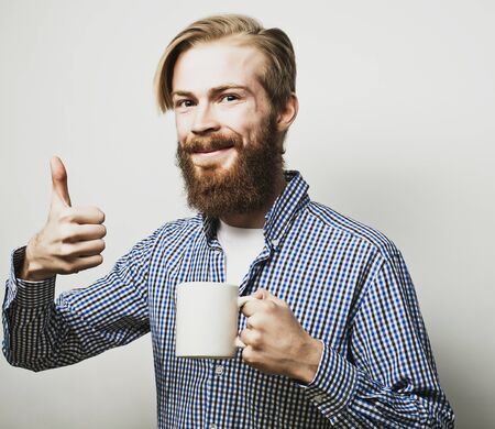 life style, happiness and people concept: young bearded man with a cup of coffee in hand and showing okey, against grey background.の写真素材