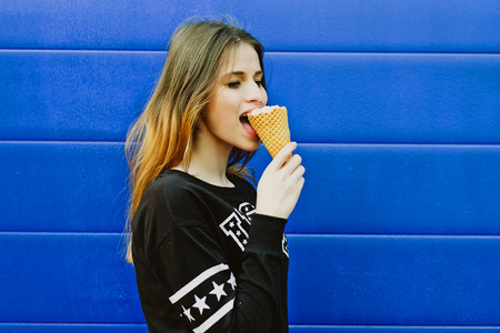 Portrait of young happy girl eating ice-cream, outdoor, over blue wall background