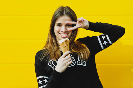 Outdoor fashion portrait of young hipster girl with  ice-cream on yellow wall backgroundの写真素材
