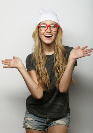 Fashion studio portrait of pretty young hipster blonde woman with glasses , wearing stylish urban t shirt and hat, over white backgroundの写真素材