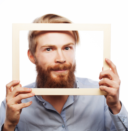 Funny picture. Young man wearing beard holding picture frame in front of his face and fun hamming, isolated on white.の写真素材