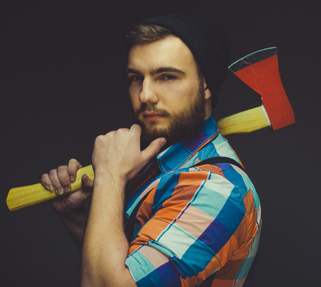 Serious lumberjack. Confident young bearded man holding a big axe and looking at camera while...