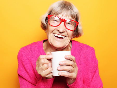 Portrait of old excited lady smiling laughing, holding cup drinking coffee, tea, beverage on yellow backgroundの写真素材