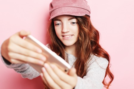 cheerful little girl with curly hair l taking a selfie isolated over pink background