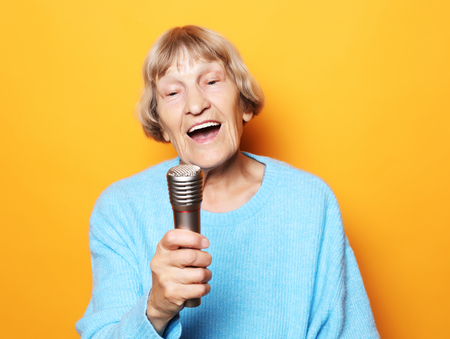 Happy old senior woman singing with microphone, having fun, expressing musical talent over yellow backgroundの写真素材