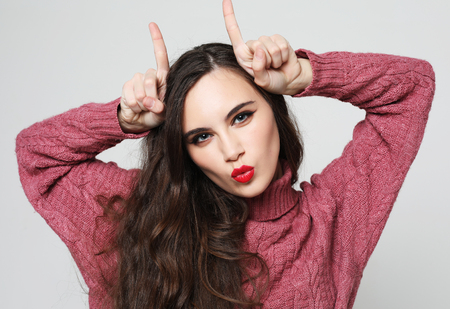 funny joking young woman, feels happy, makes horns on her head with fingers, wears pink sweaterの写真素材