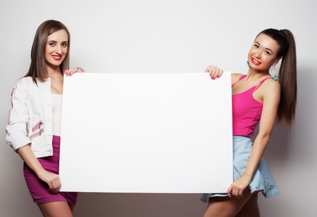 lifestyle, emotion and people concept: Two young girl friends standing together and holding a blank billboardの写真素材