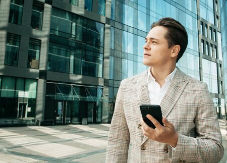 young businessman dressed in formal clothes standing outside a glass building and using mobileの写真素材