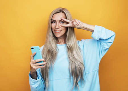 Smiley senior woman is holding a new smartphone in her hand over yellow background