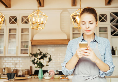 A beautiful young woman in the kitchen holds a smartphone and types a message.の写真素材