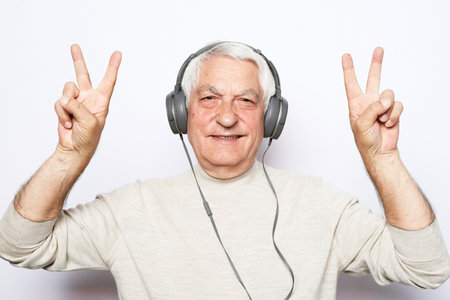 Lifestyle, tehnology and old people concept: Handsome old man in casual wear and headphones is listening to music and show victory sign on white backgroundの写真素材