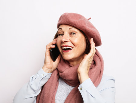 Cheerful successful old woman wearing pink scarf and beret and talking on the phone. Positive emotions. Lifestyle concept. Close up over white background.の写真素材