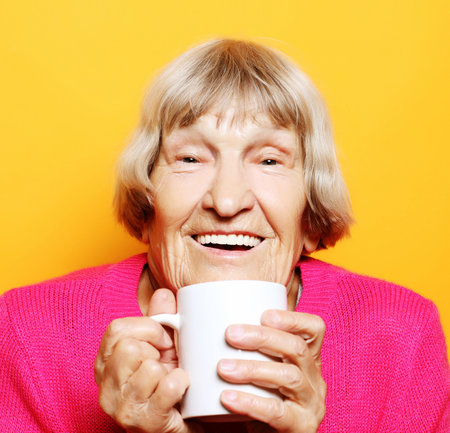 old excited lady smiling laughing, holding cup drinking coffee, tea, beverage on yellow background